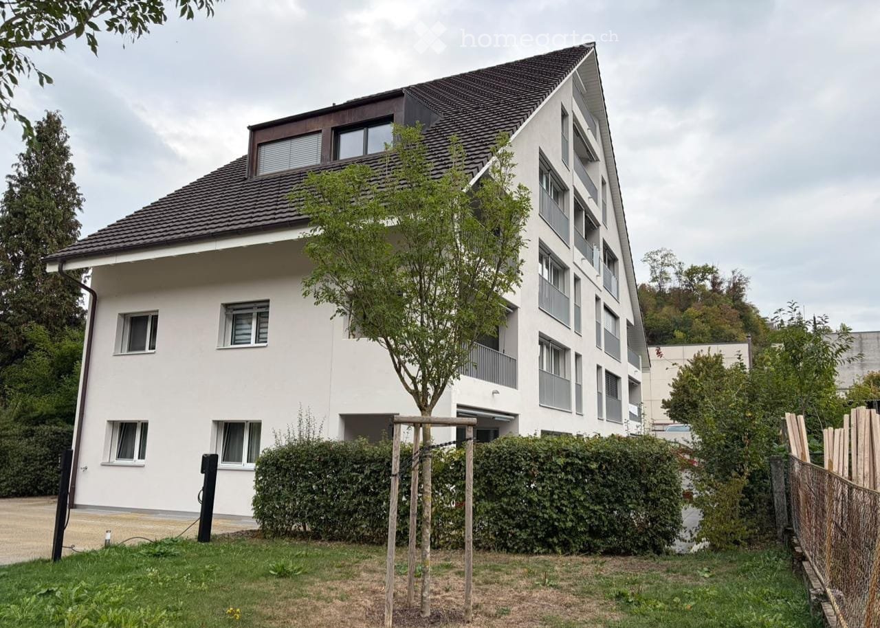 Modern two-story residential building with white exterior, multiple windows, and a sloped roof. Features a small garden area with a tree and wooden play structure in the foreground.