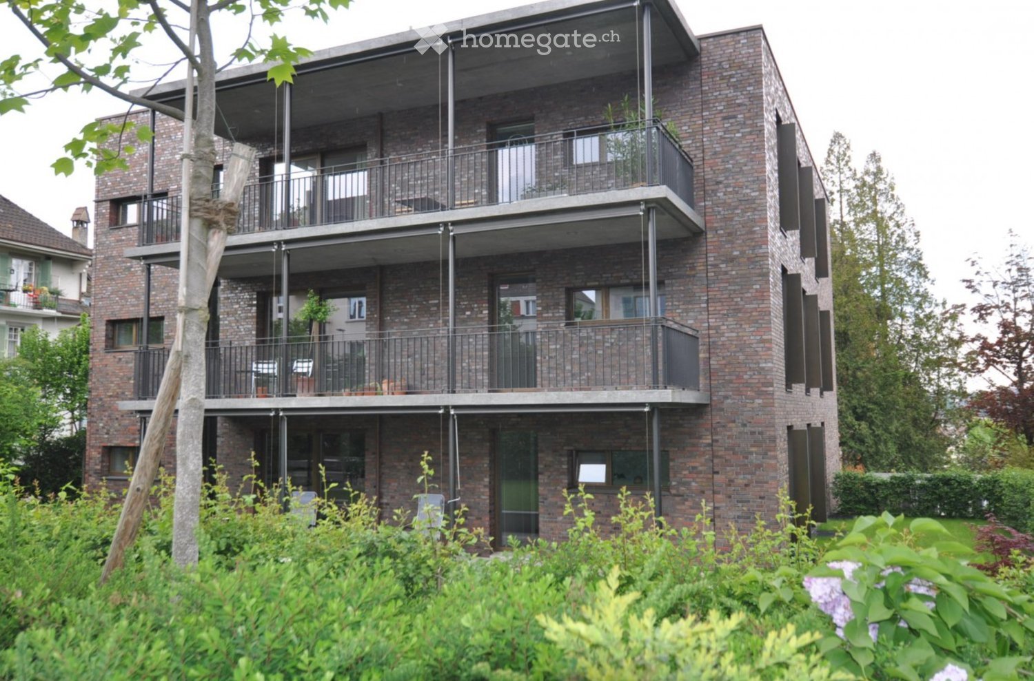 Modern multi-story brick building with balconies and large windows, surrounded by greenery.