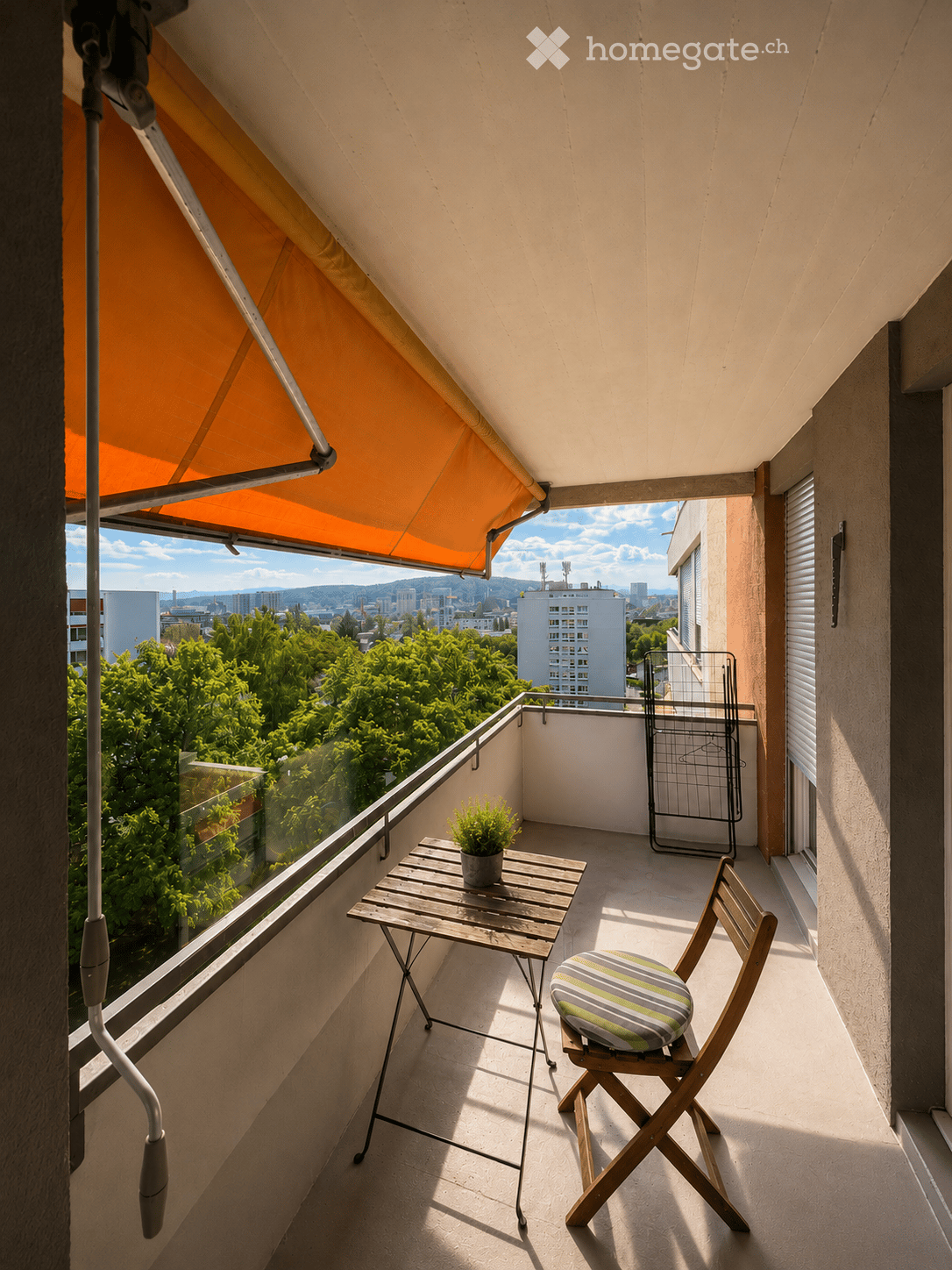 Balcony with a wooden table, chair, and small potted plant, featuring a retractable awning and a view of greenery and cityscape.