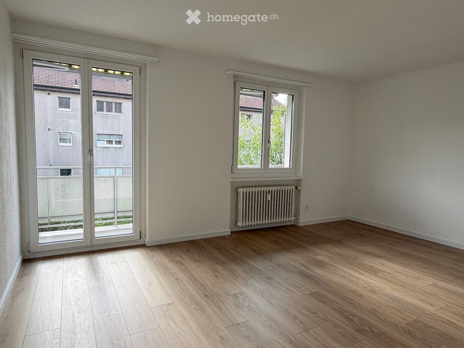 Empty room with wooden flooring, large sliding glass door leading to a balcony, and a window with radiator below.