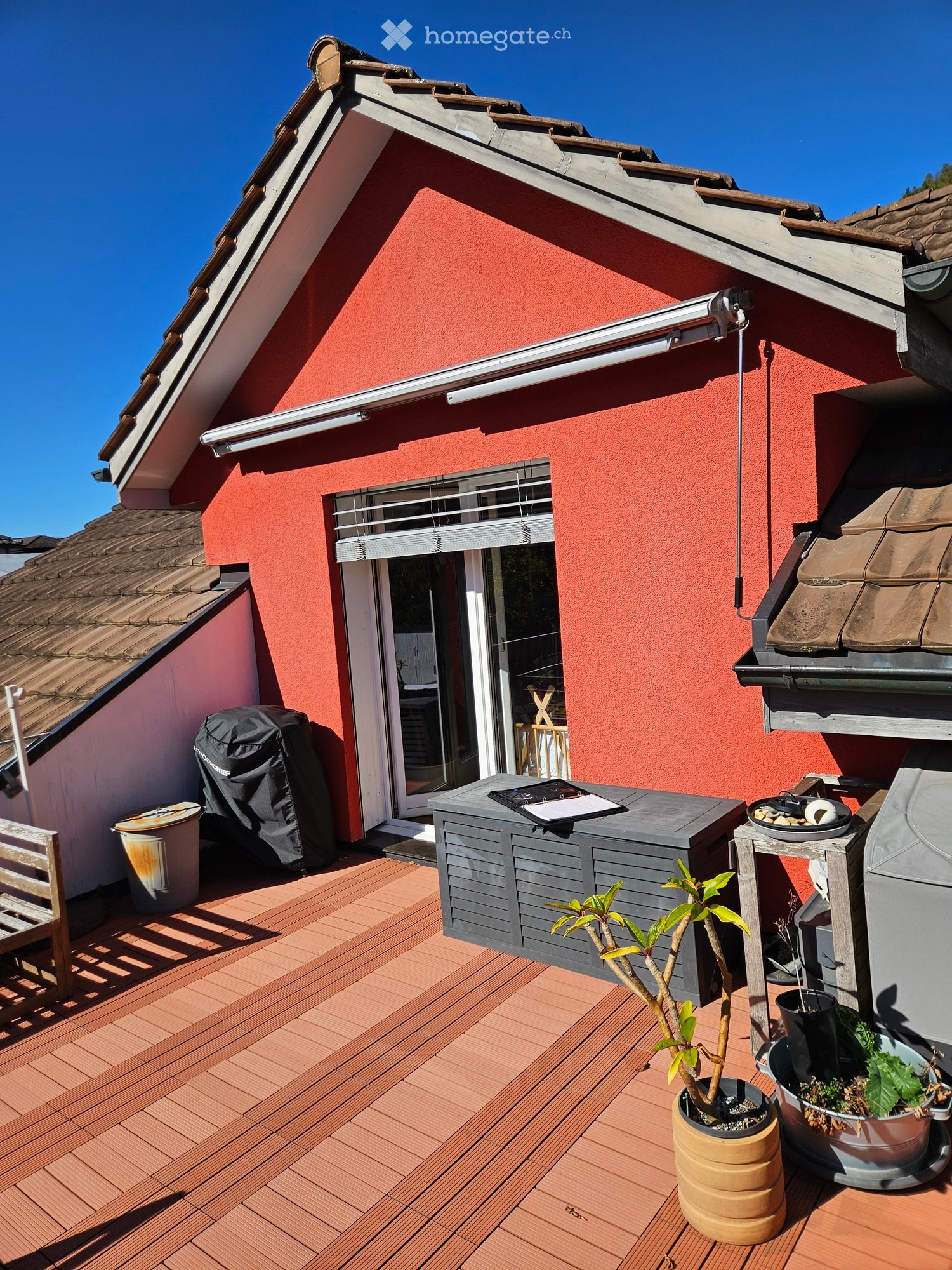 Wooden deck with a grey table, chairs, potted plants, and a covered grill. The deck is attached to a red house with a sliding glass door.