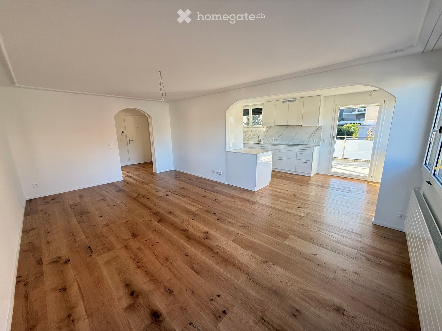 Empty room with light wooden flooring, white walls, and a white kitchen area with cabinets and a countertop visible in the background.