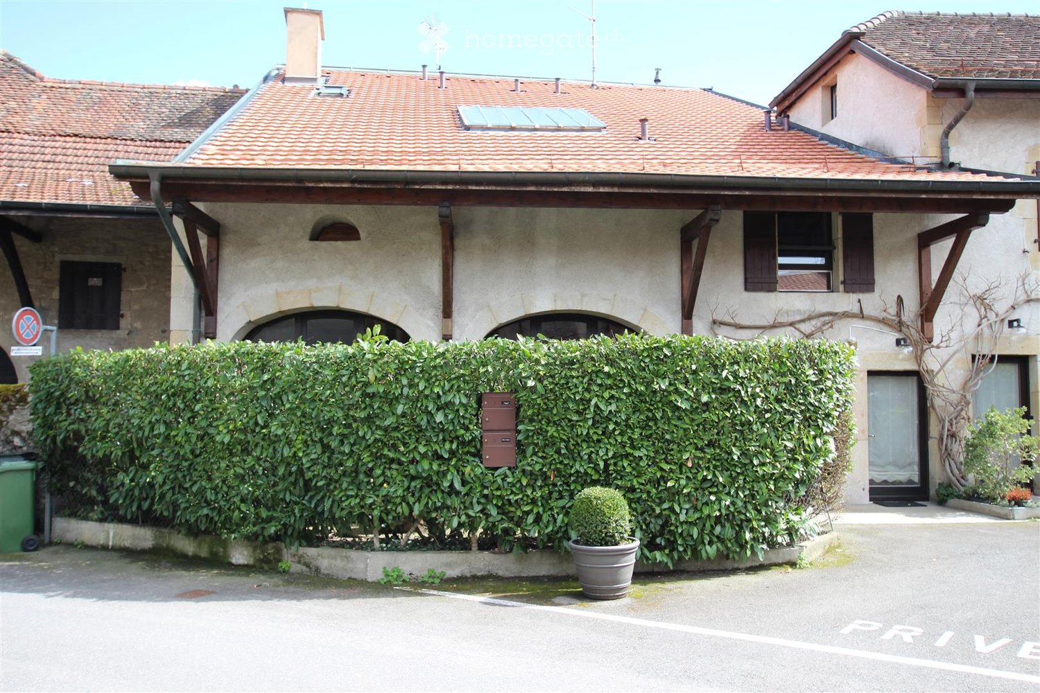 Two-story residential building with a tiled roof, stone facade, and wooden shutters. The entrance is partially obscured by a tall hedge, with a small potted plant and a green trash bin visible.