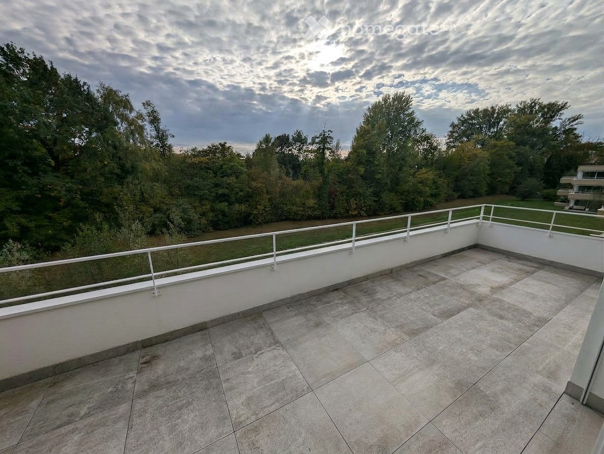 Empty balcony with tiled floor, white railing, and a view of a green area with trees and a body of water.