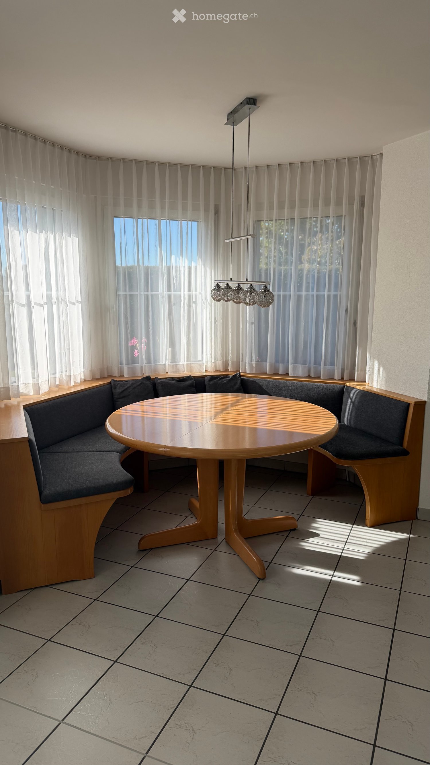 A modern dining area with a round wooden table, built-in bench seating, large windows with sheer curtains, and a hanging light fixture.