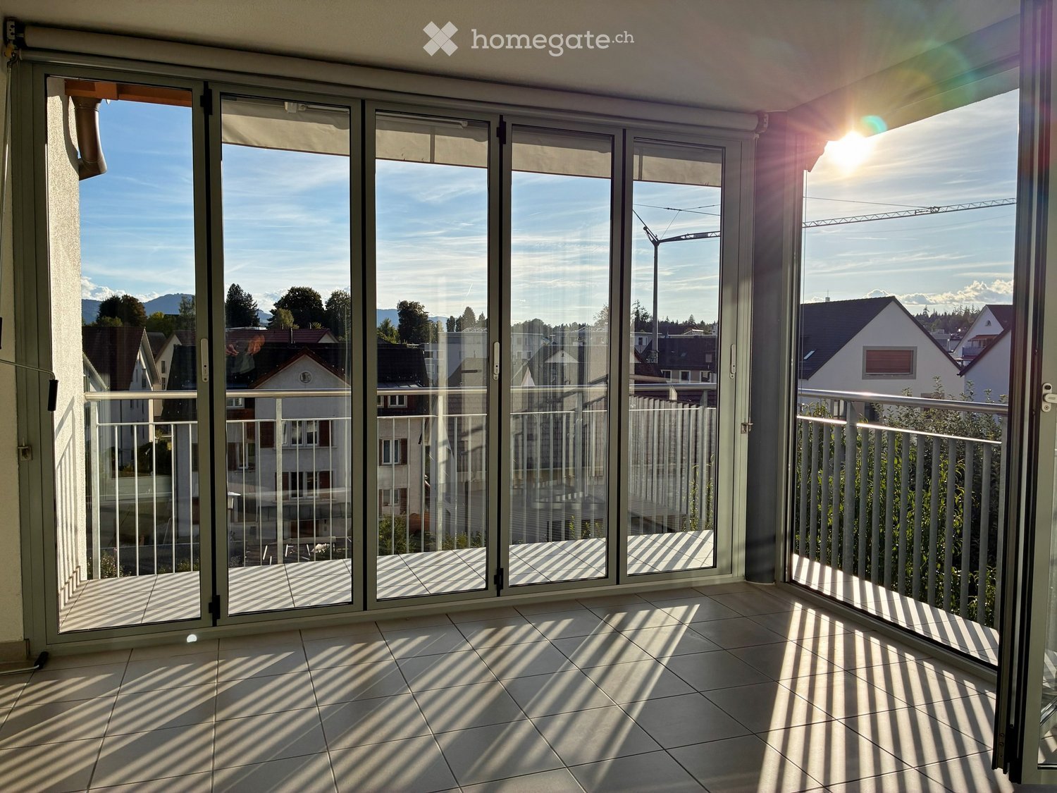 Spacious balcony with large glass sliding doors, tiled floor, and metal railing, offering a view of the neighborhood and mountains.