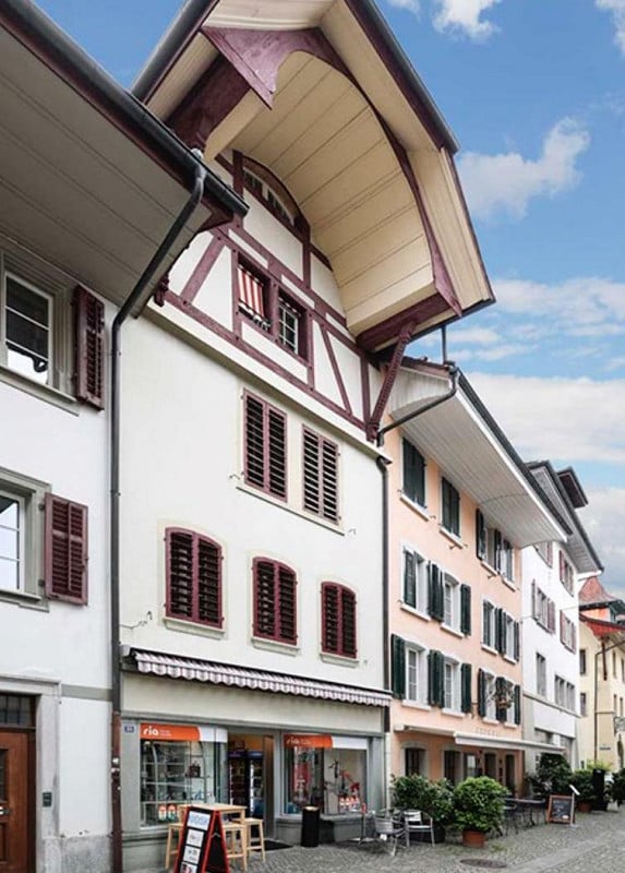 A multi-story building with a distinctive half-timbered upper level, white walls, and red shutters. The ground floor features a storefront with a striped awning and a few outdoor tables and chairs.
