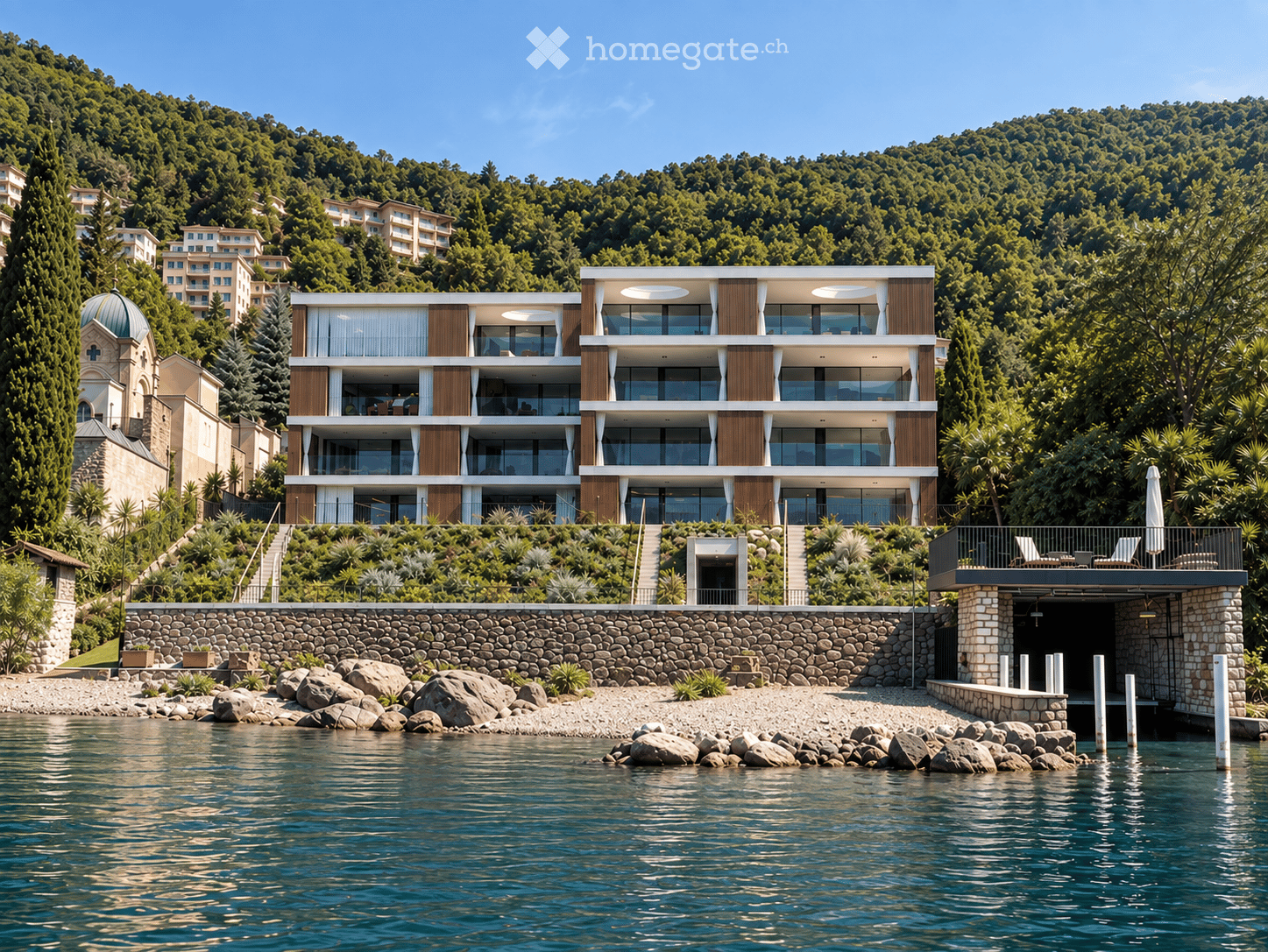 Modern multi-story building with large windows and balconies, situated on a hillside overlooking a body of water, surrounded by greenery and other structures.