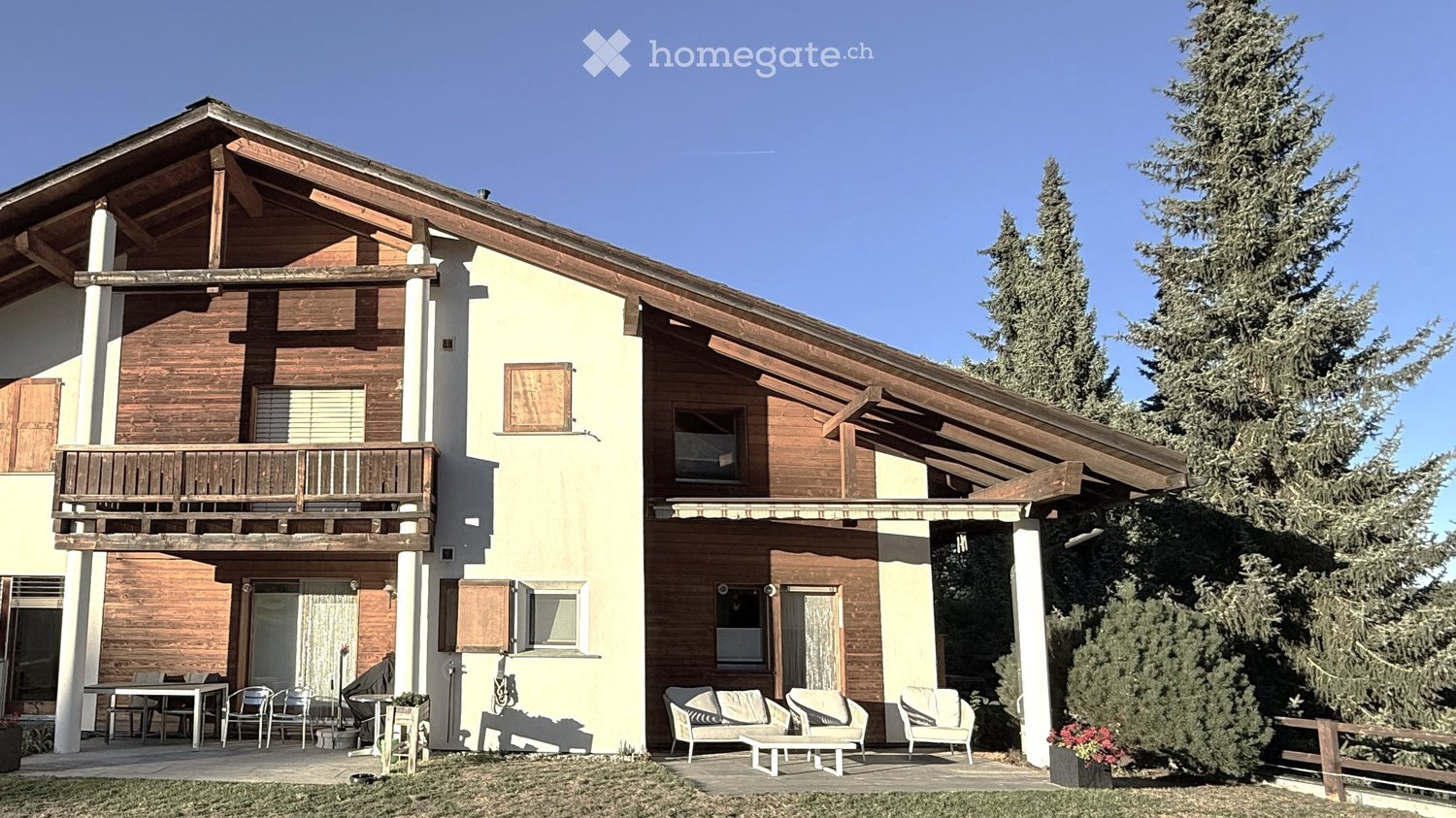 Two-story house with wooden and white exterior, balcony, patio seating area, and surrounding greenery.