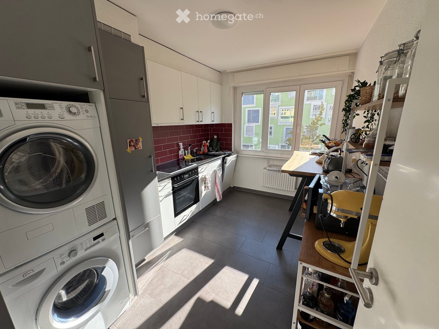 Modern kitchen with white cabinets, dark tiled floor, and a window overlooking a green building. Includes a washing machine, dishwasher, oven, sink, and a small table with a yellow mixer.
