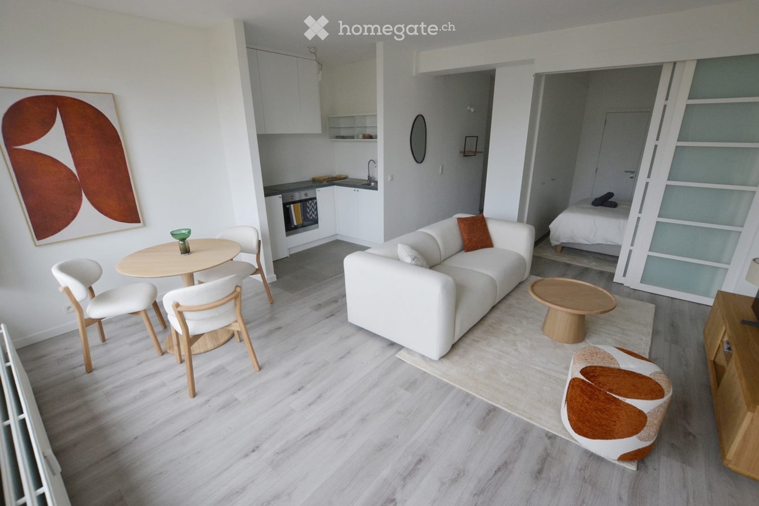Modern, open-concept living space with a white sofa, coffee table, dining area, and a separate bedroom with a bed. The kitchen features white cabinets and stainless steel appliances.