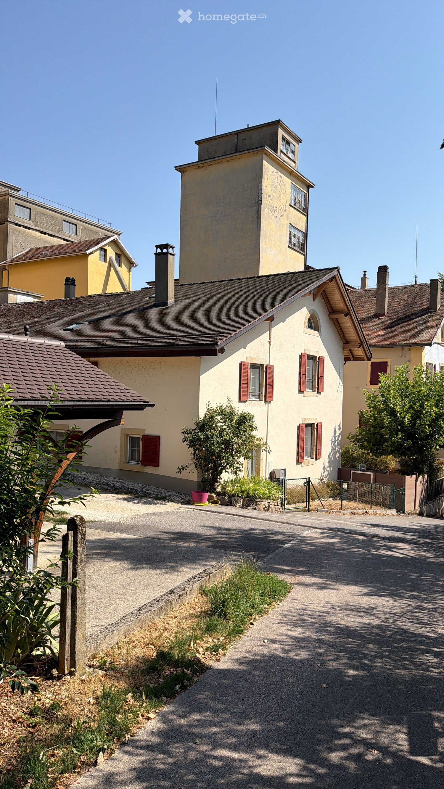 A two-story residential building with beige walls, red shutters, and a tiled roof, surrounded by greenery and a paved driveway.