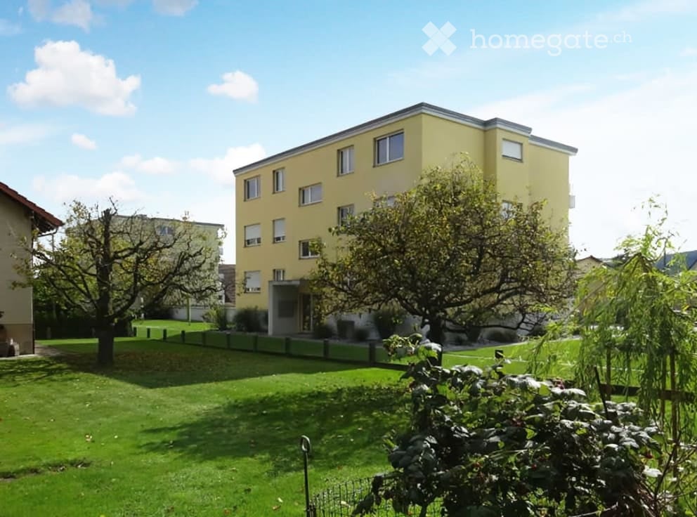 Apartment building with a yellow facade, multiple windows, fenced lawn, and trees in the yard