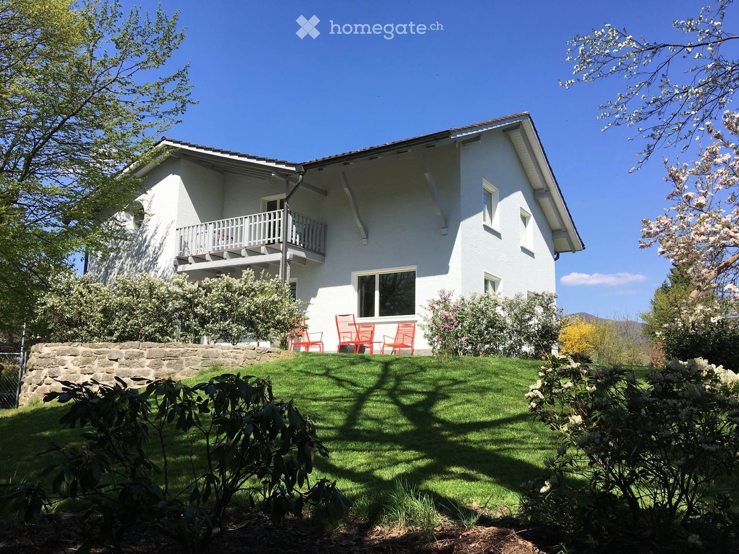 A two-story white house with a balcony, surrounded by a well-maintained lawn and flowering shrubs.