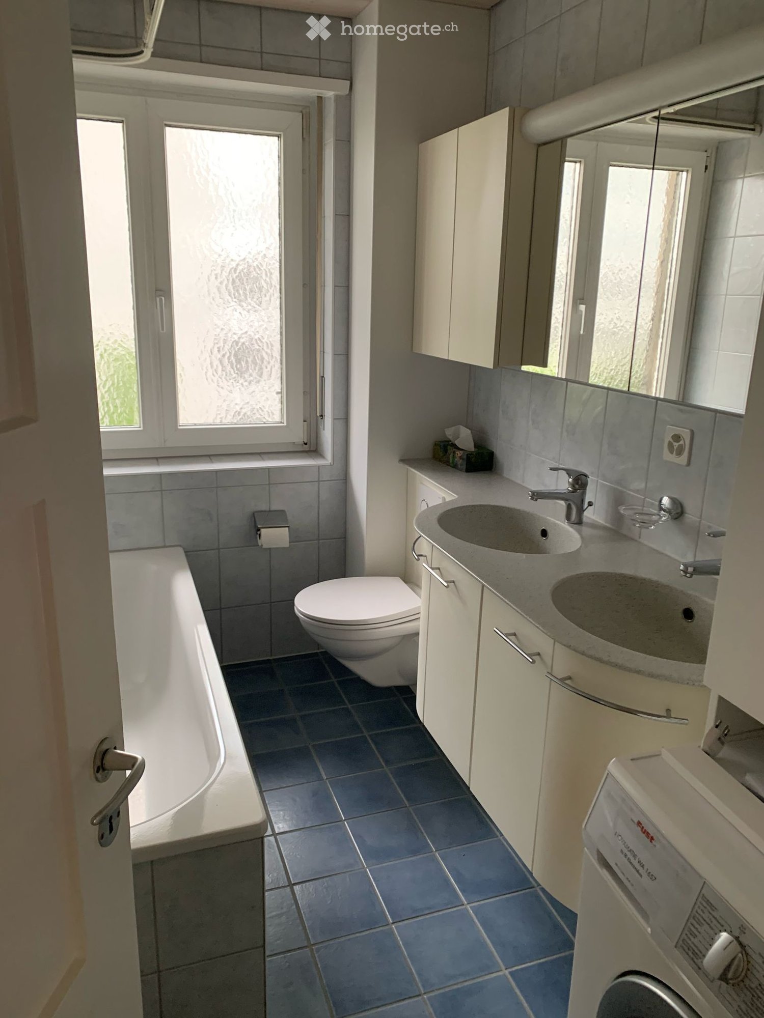 Modern bathroom with blue tiled floor, white bathtub, toilet, double sink with modern faucet, mirrored cabinet, and a washing machine in the corner.