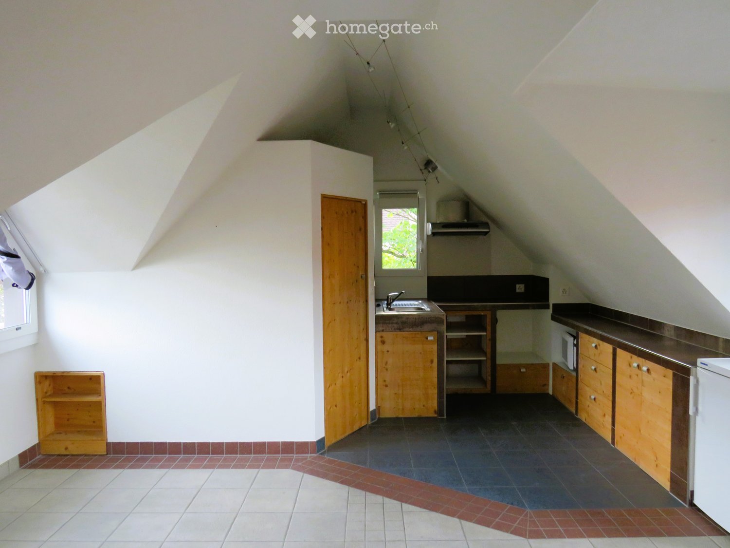 Bright attic kitchen with wooden cabinets, dark countertops, and a sink. Features include a window, shelves, and a refrigerator.