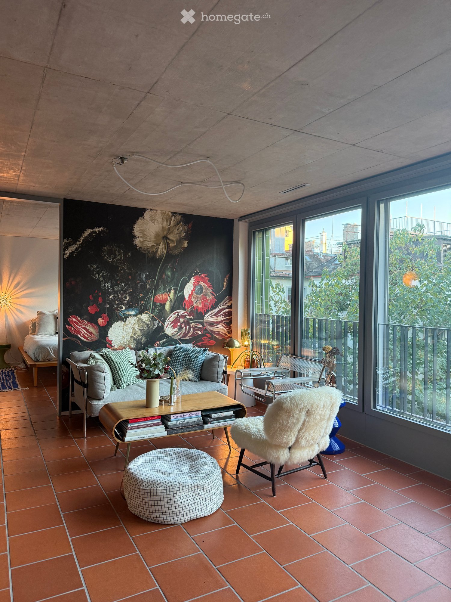 Modern living room with terracotta tiles, large windows, and a view of greenery. Features a grey sofa, coffee table with books and a vase, ottoman, fur chair, and a bed in the background.