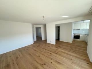 Empty room with wooden flooring and white walls, featuring a doorway leading to another room and a kitchen area with white cabinets and a countertop.