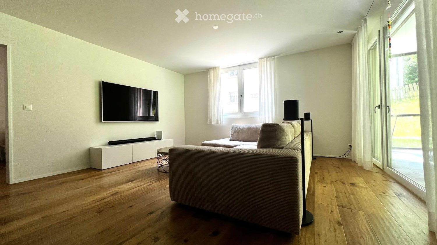 Modern living room with light-colored walls, wooden flooring, a grey sofa, a coffee table, a television mounted on the wall, and a floor lamp near the window.
