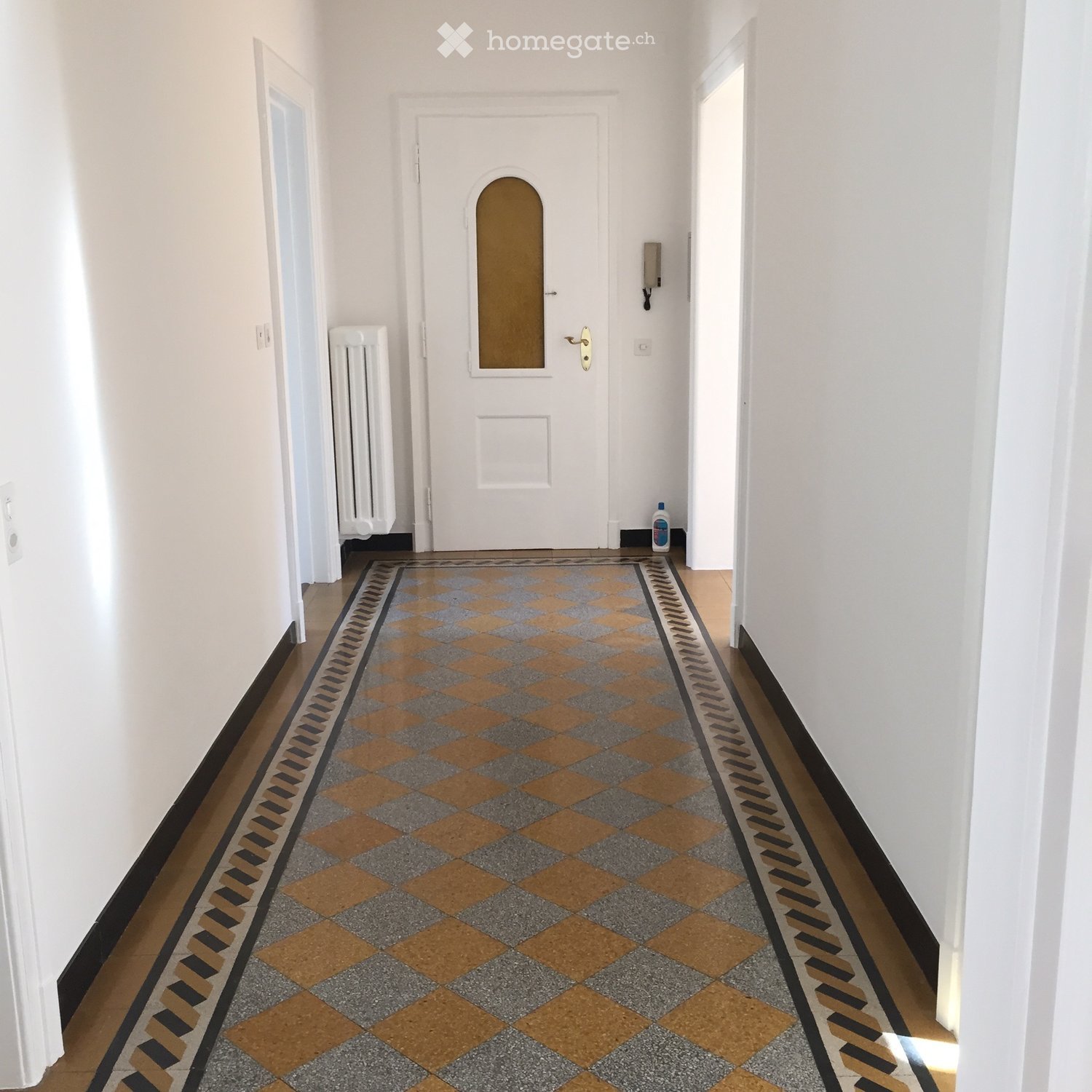 A long, narrow hallway with white walls, a checkered floor in orange and gray, and a central white door with a decorative glass panel. A radiator is visible on the left wall, and a small cleaning bottle sits on the floor near the right wall.