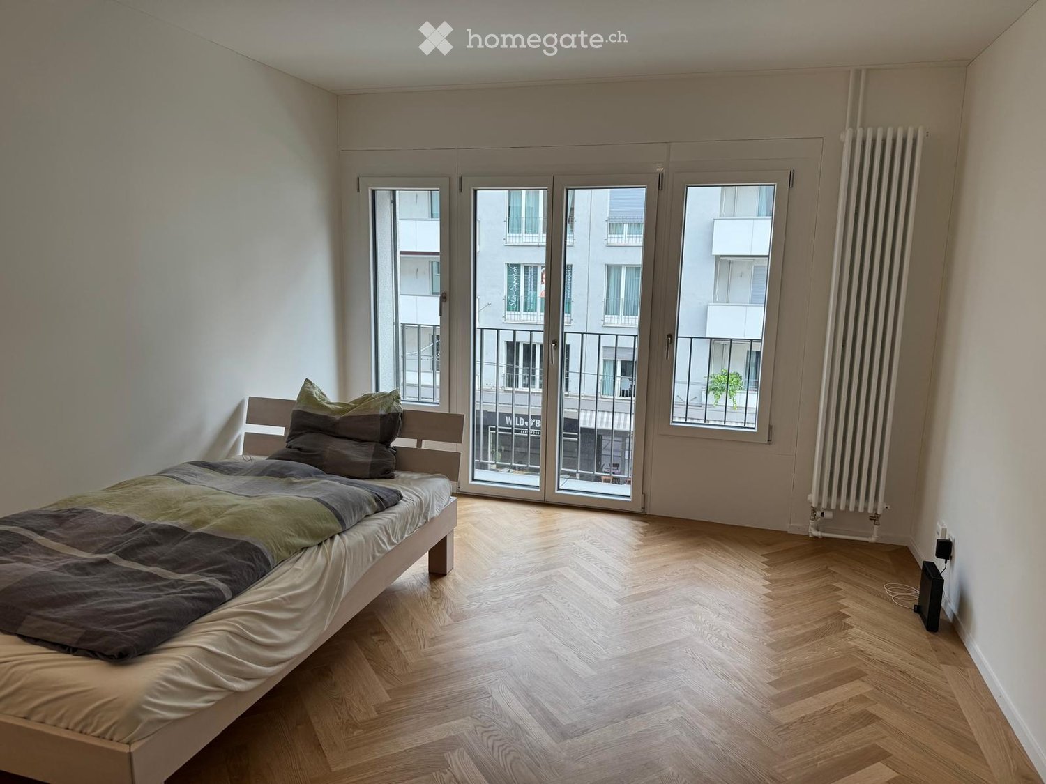 Minimalist bedroom with a single bed, wooden flooring, large windows with sliding doors leading to a balcony, and a radiator on the wall.