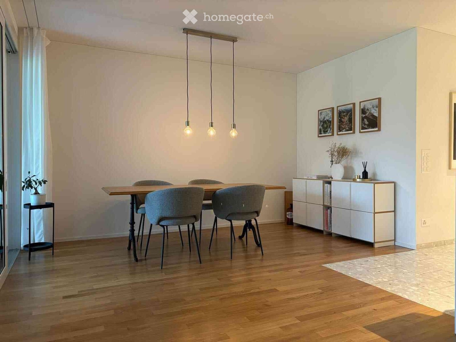 large dining table with chairs, wooden floor, white walls, hanging lamps, cabinet with books, potted plant