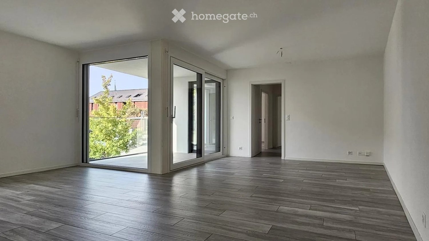 Empty room with wooden flooring, large sliding glass doors leading to an outdoor area, and a doorway to another room.