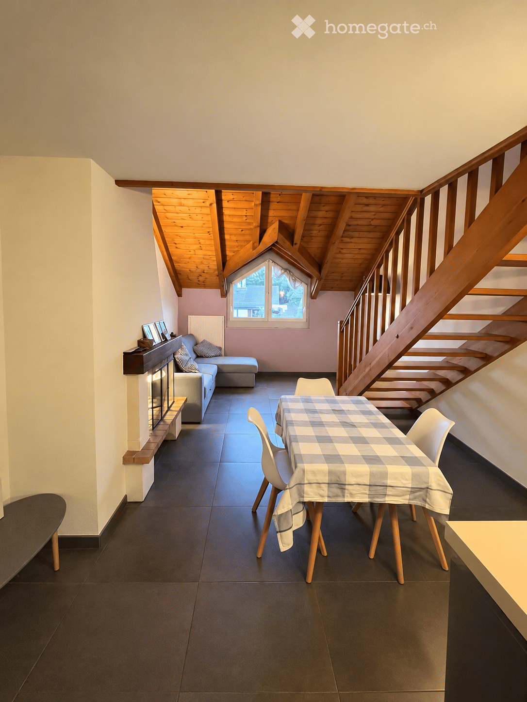 Bright living room with a wooden slanted ceiling, dark tiled floor, and a cozy seating area featuring a sofa, ottoman, and fireplace. A dining table with chairs is set with a checkered tablecloth, and a staircase leads upwards.