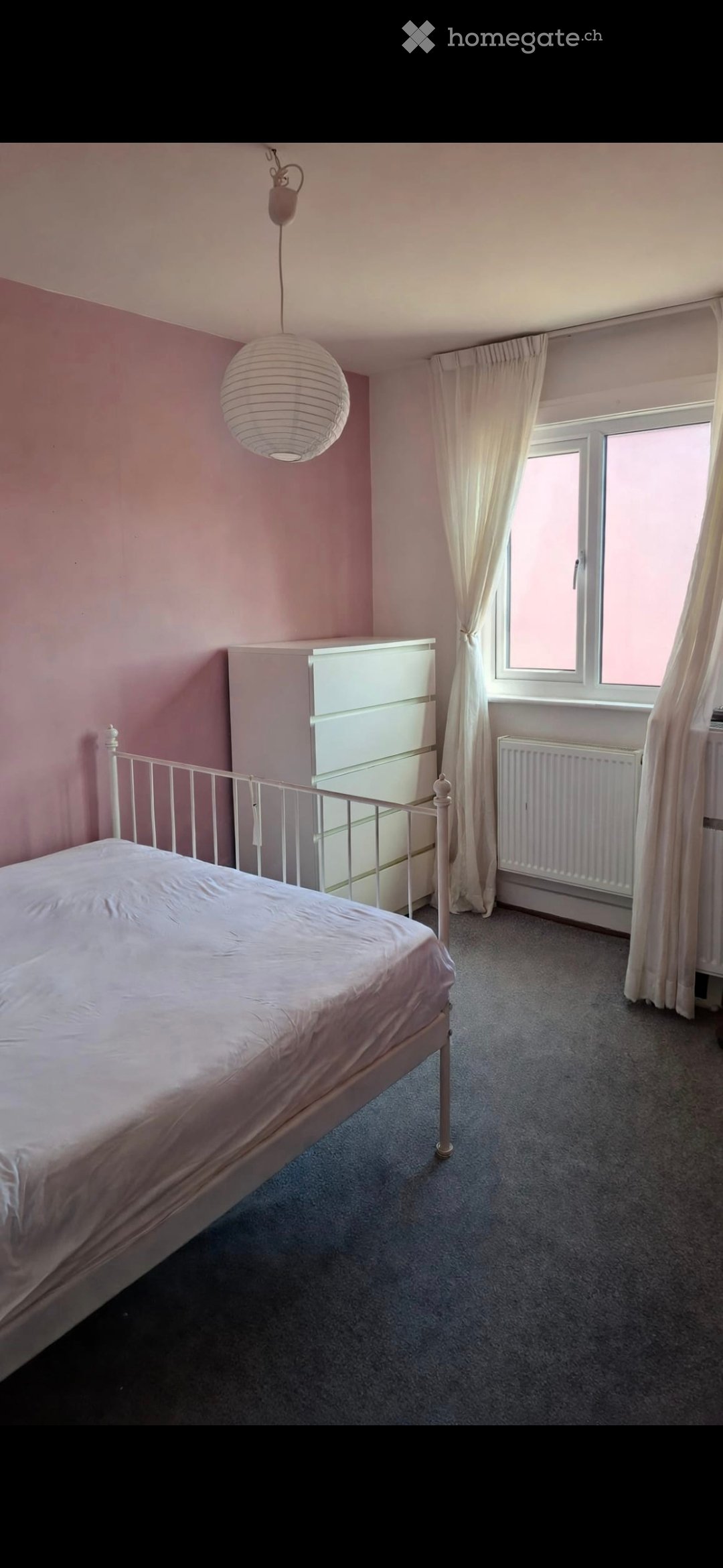 A furnished bedroom with a single bed, white wardrobe, radiator, and a hanging paper lantern light fixture. The room features light pink walls, grey carpet, and white curtains on the window.