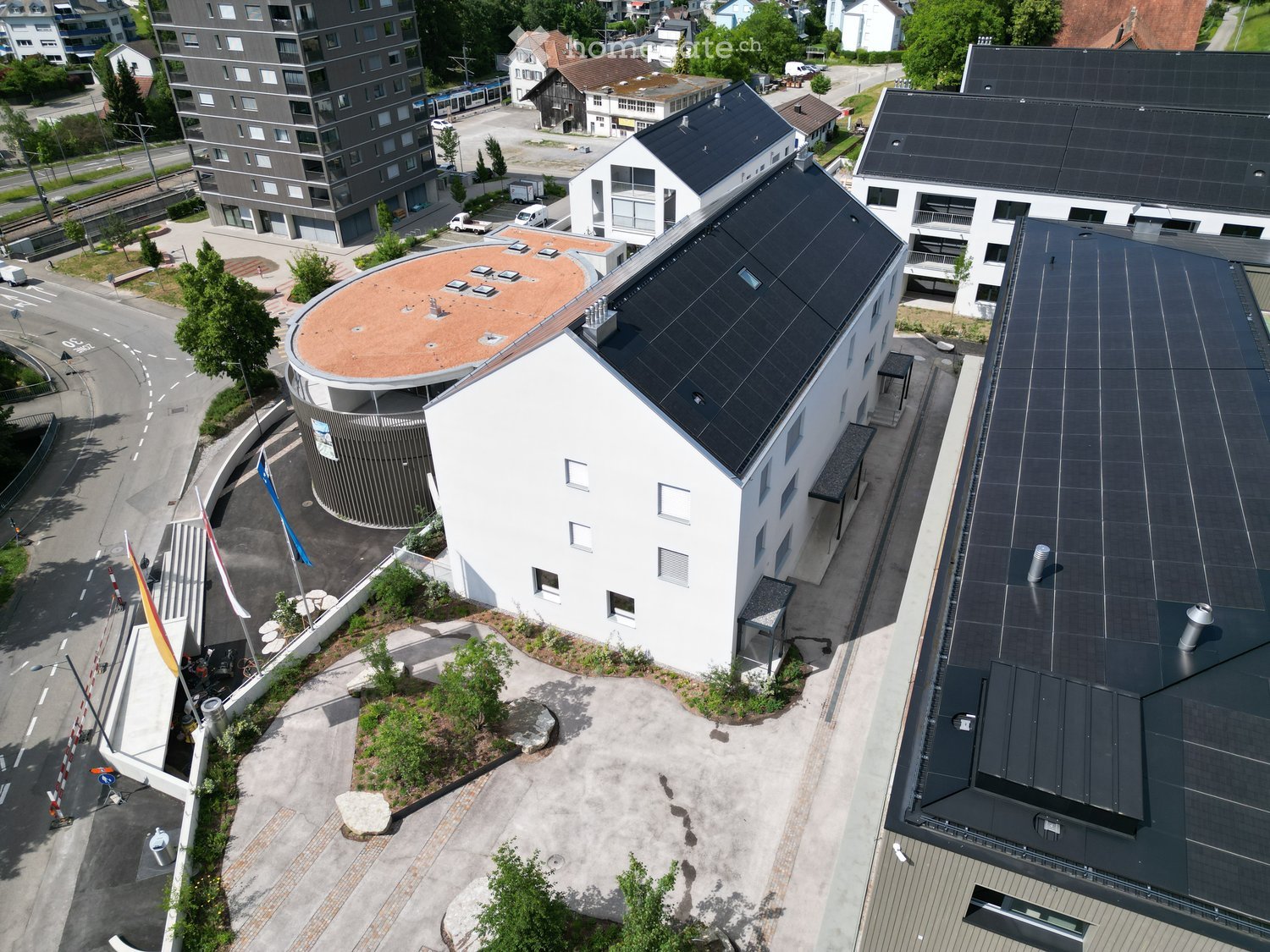 Aerial view of a modern multi-story building complex with solar panels on the roof, surrounded by other structures and greenery.