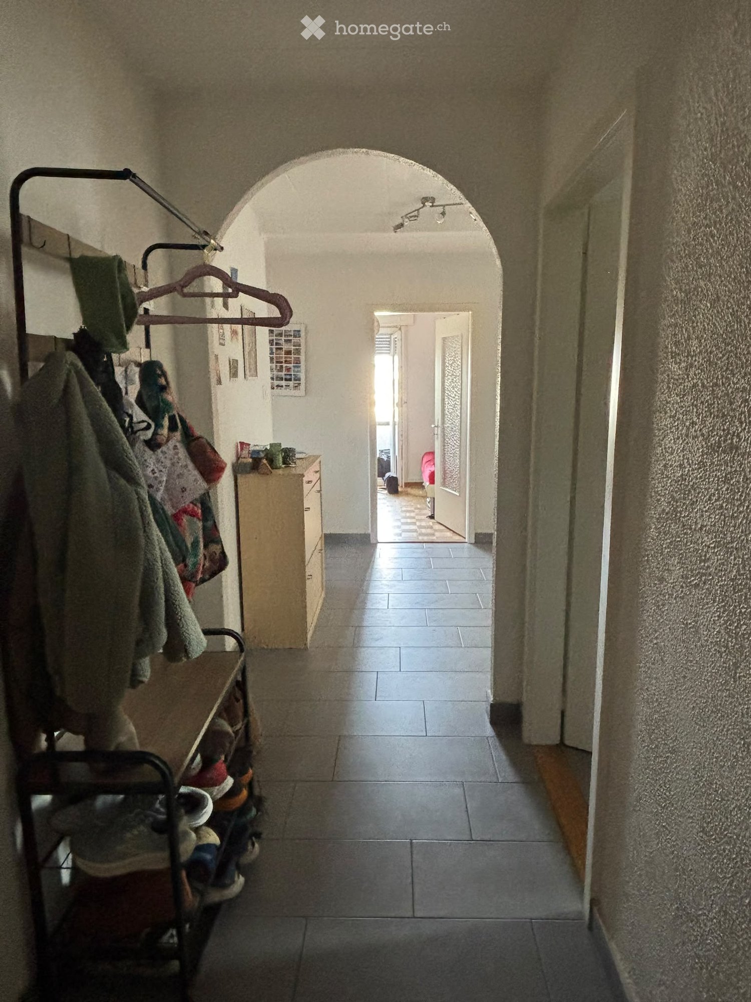A narrow hallway with tiled flooring, arched ceiling, and a coat rack on the left side. The hallway leads to an open doorway at the end, with a small table and some items visible in the background.