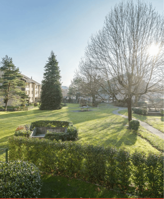 Well-maintained garden with neatly trimmed hedges, a central lawn, a small pond, and a few trees providing shade. The area is bordered by a low fence and features a winding pathway.