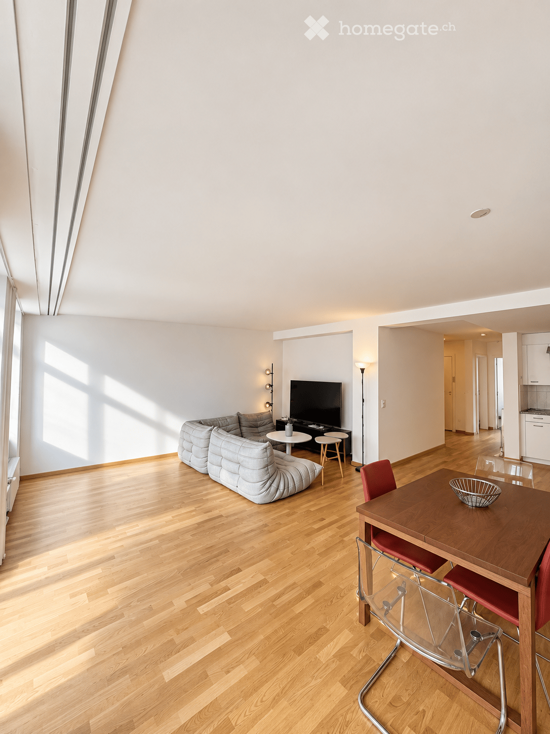 Open-plan living and dining area with light wooden flooring, a grey sectional sofa, a coffee table, a TV, a dining table with chairs, and a floor lamp.