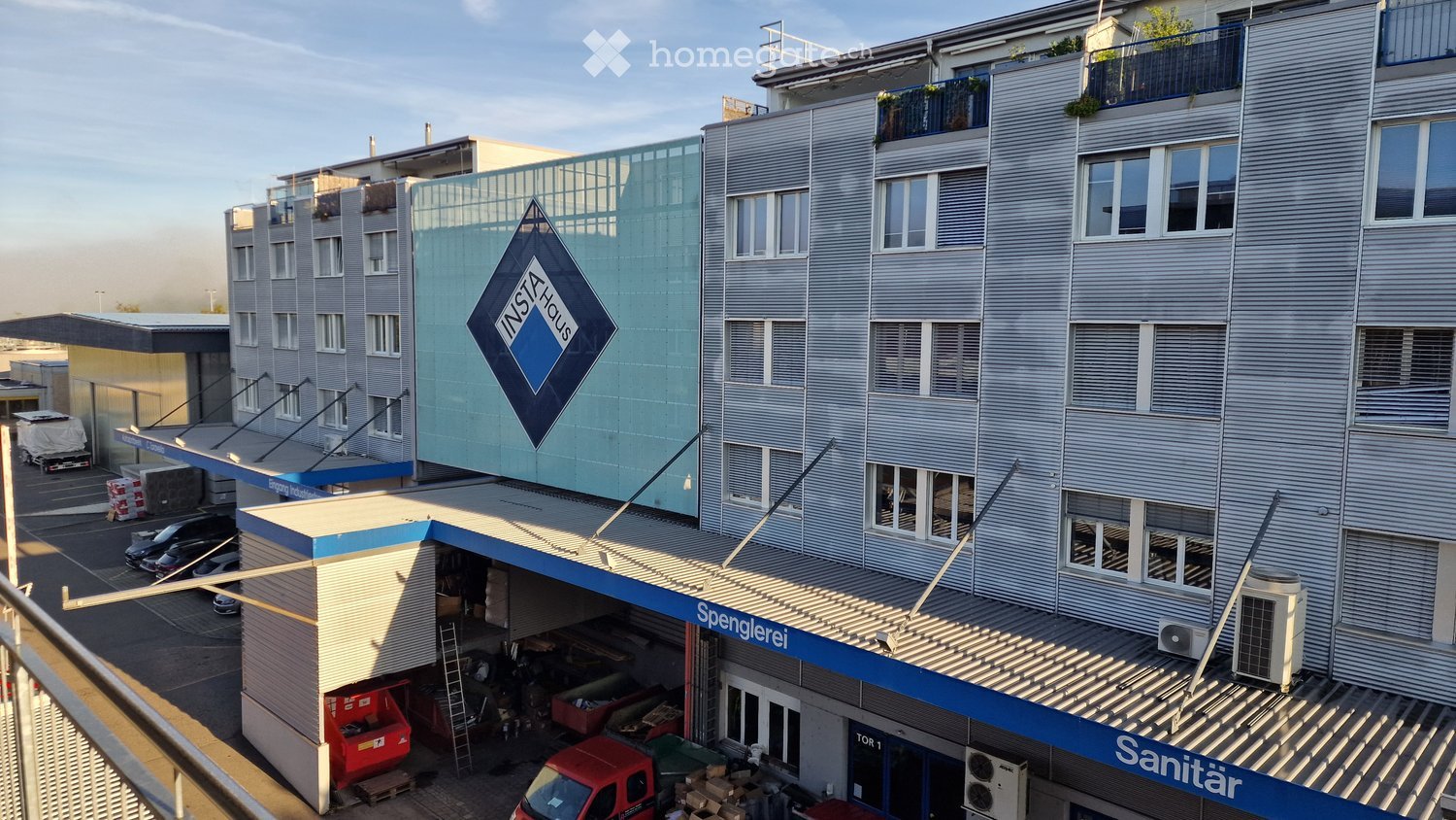 Multi-story commercial building with a garage, parking area, and signage for "Spenglerei" and "Sanitär".