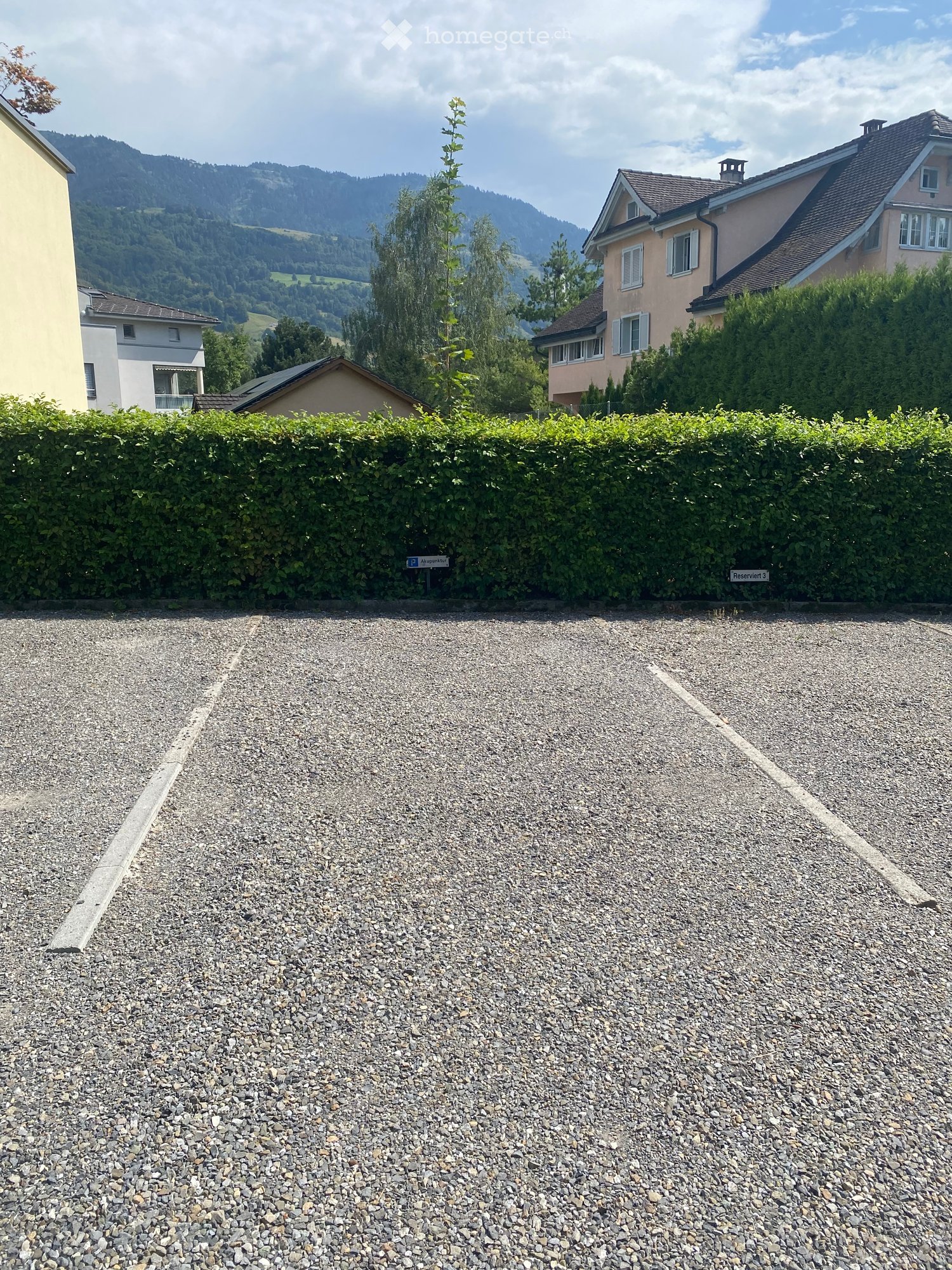 Gravel parking area in front of a hedge with houses and mountains in the background.