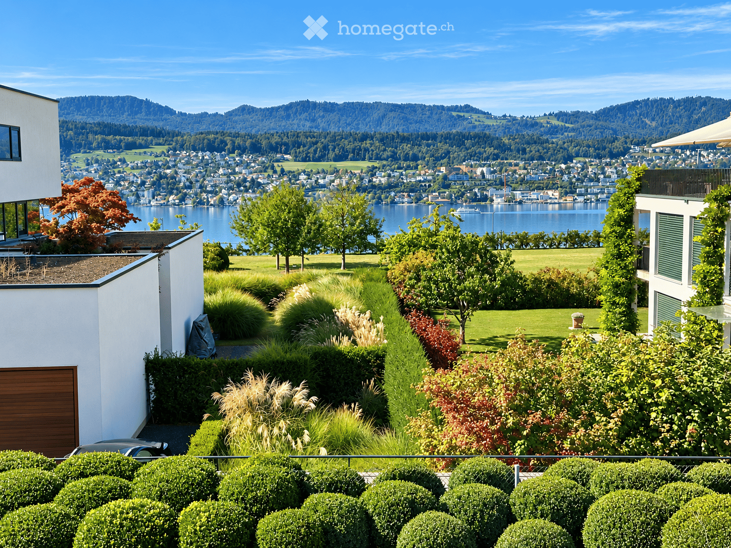 A well-maintained garden with neatly trimmed hedges, various plants, and trees, overlooking a scenic lake and distant mountains.