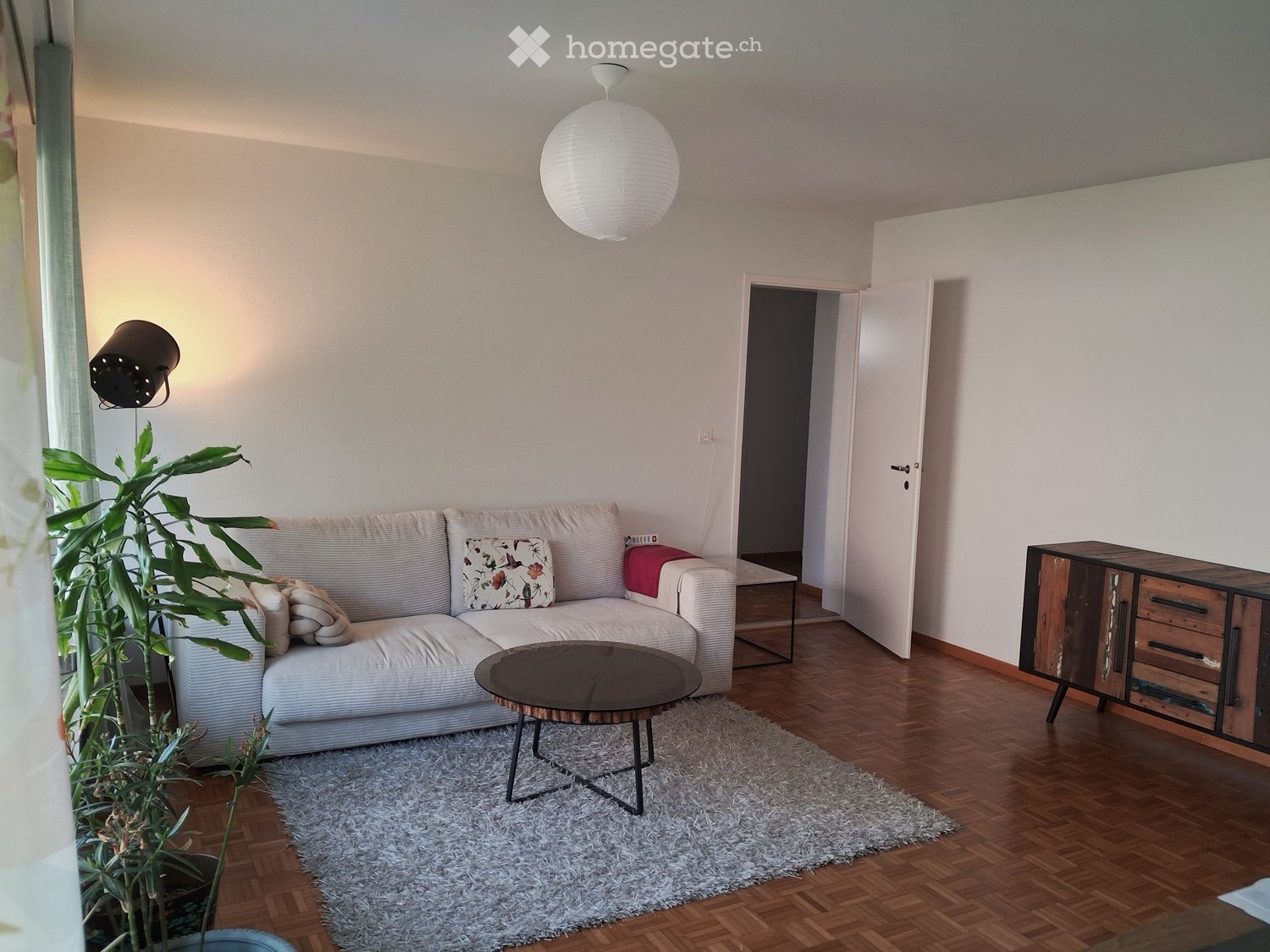 Modern living room with a light grey sofa, wooden coffee table, side table, storage cabinet, floor lamp, and a potted plant. The room features wooden flooring and white walls.