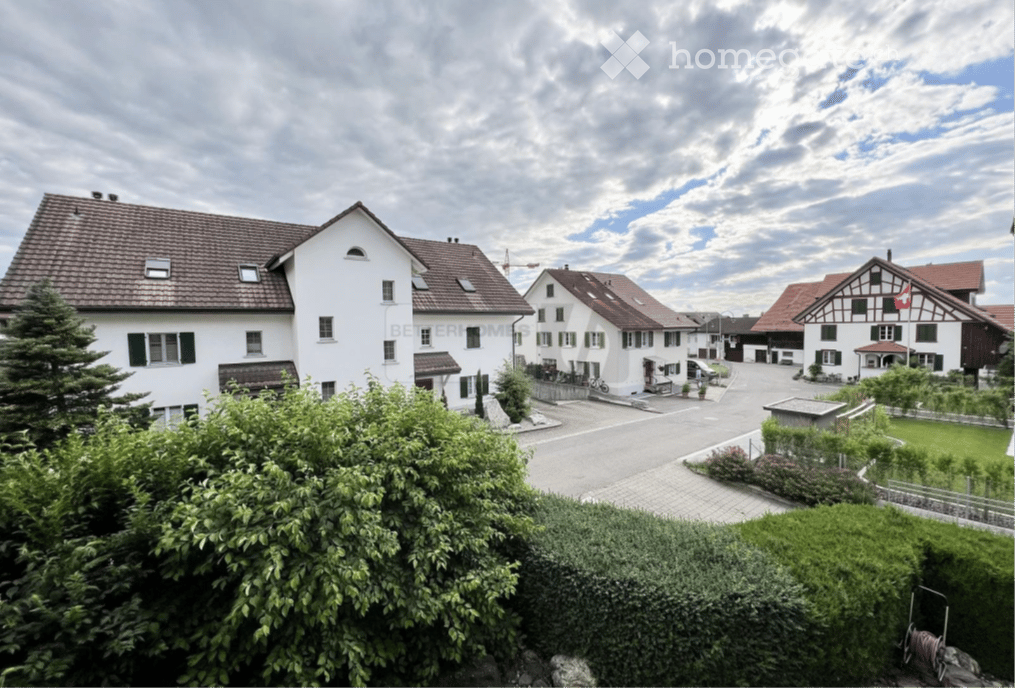 A row of traditional Swiss houses with pitched roofs and white facades, surrounded by greenery and a paved road.