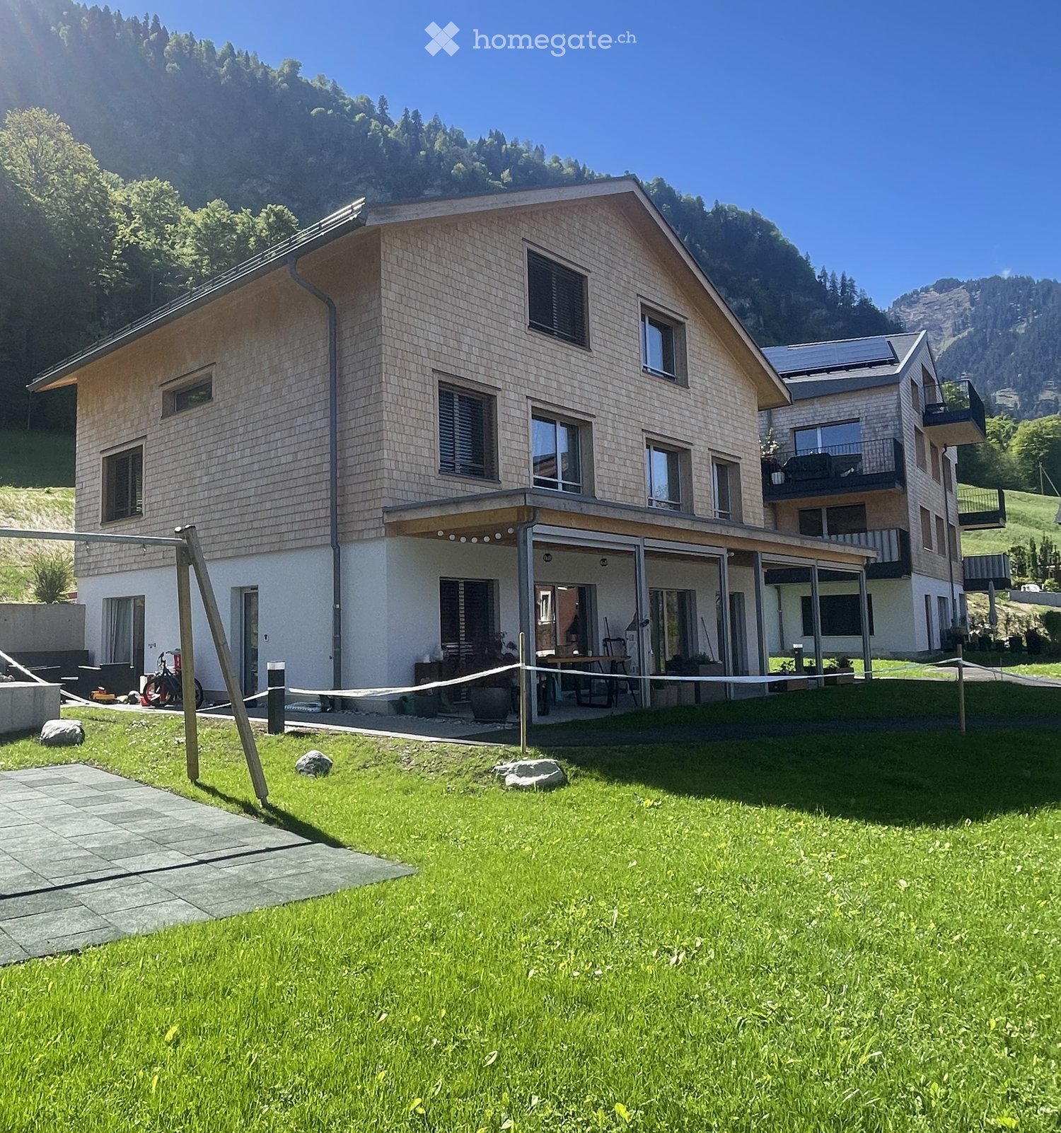Two-story modern house with light-colored siding, large windows, and a spacious covered patio area. The house is surrounded by a well-maintained lawn and is situated in a scenic, mountainous area.