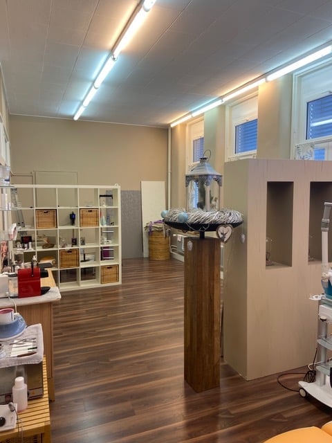 Bright, open room with wooden flooring, white shelving unit, small table with items, decorative wooden stand with a vase, and a lamp on a pedestal.