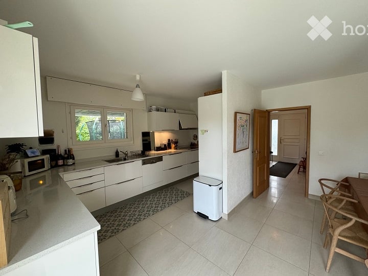 Modern kitchen with white cabinets, stainless steel appliances, and a light-colored tiled floor. Features a sink, fridge, microwave, and a small dining area with chairs and a table.