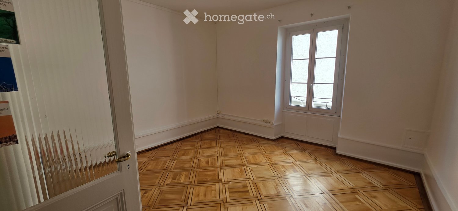 Empty room with wooden parquet flooring, white walls, and a single window with white frame. No furniture or equipment present.