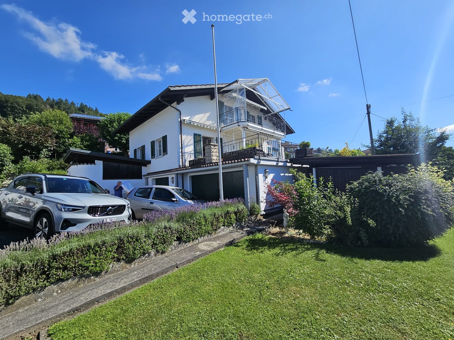Two-story house with white exterior, dark roof, garage, and balcony on the upper level. Two cars are parked in front, and the garden features well-maintained grass and shrubs.