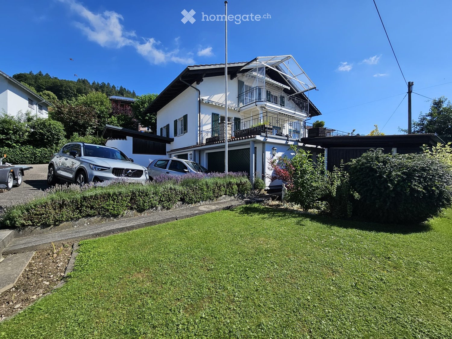 Two-story white house with a balcony, garage, and well-maintained garden with green lawn and shrubs.