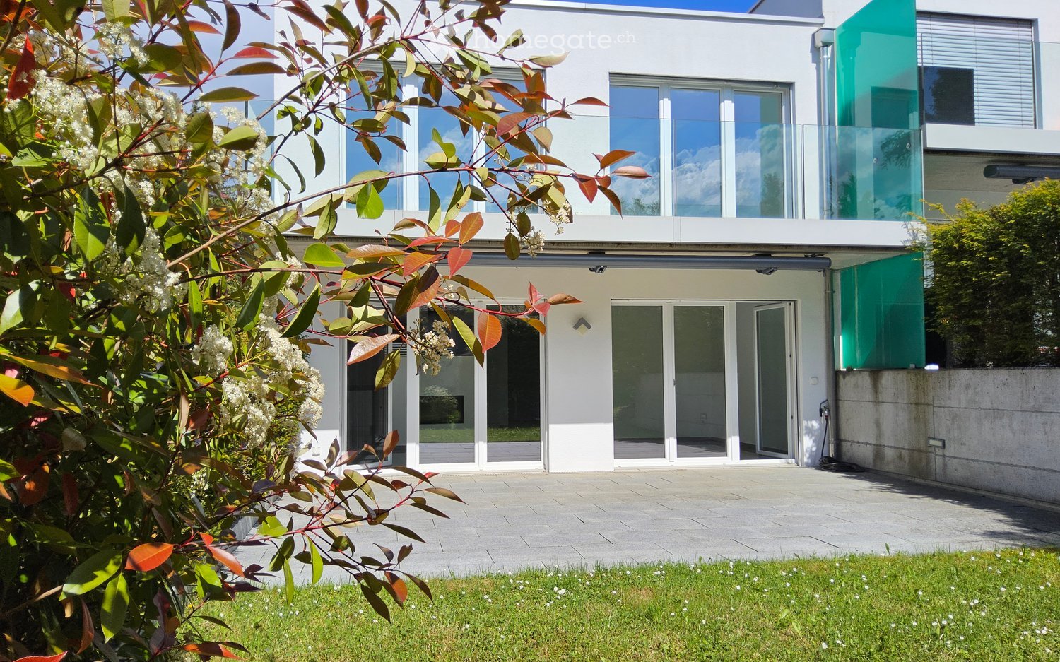 Modern two-story house with large windows, glass balcony, and a well-maintained garden area.