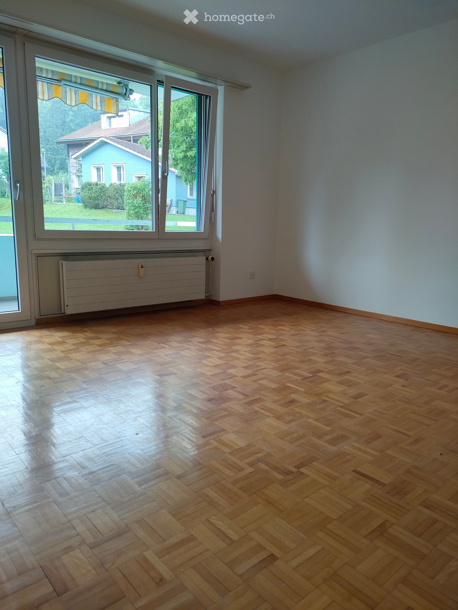 Empty room with wooden herringbone flooring, large window with blinds, radiator, and white walls.