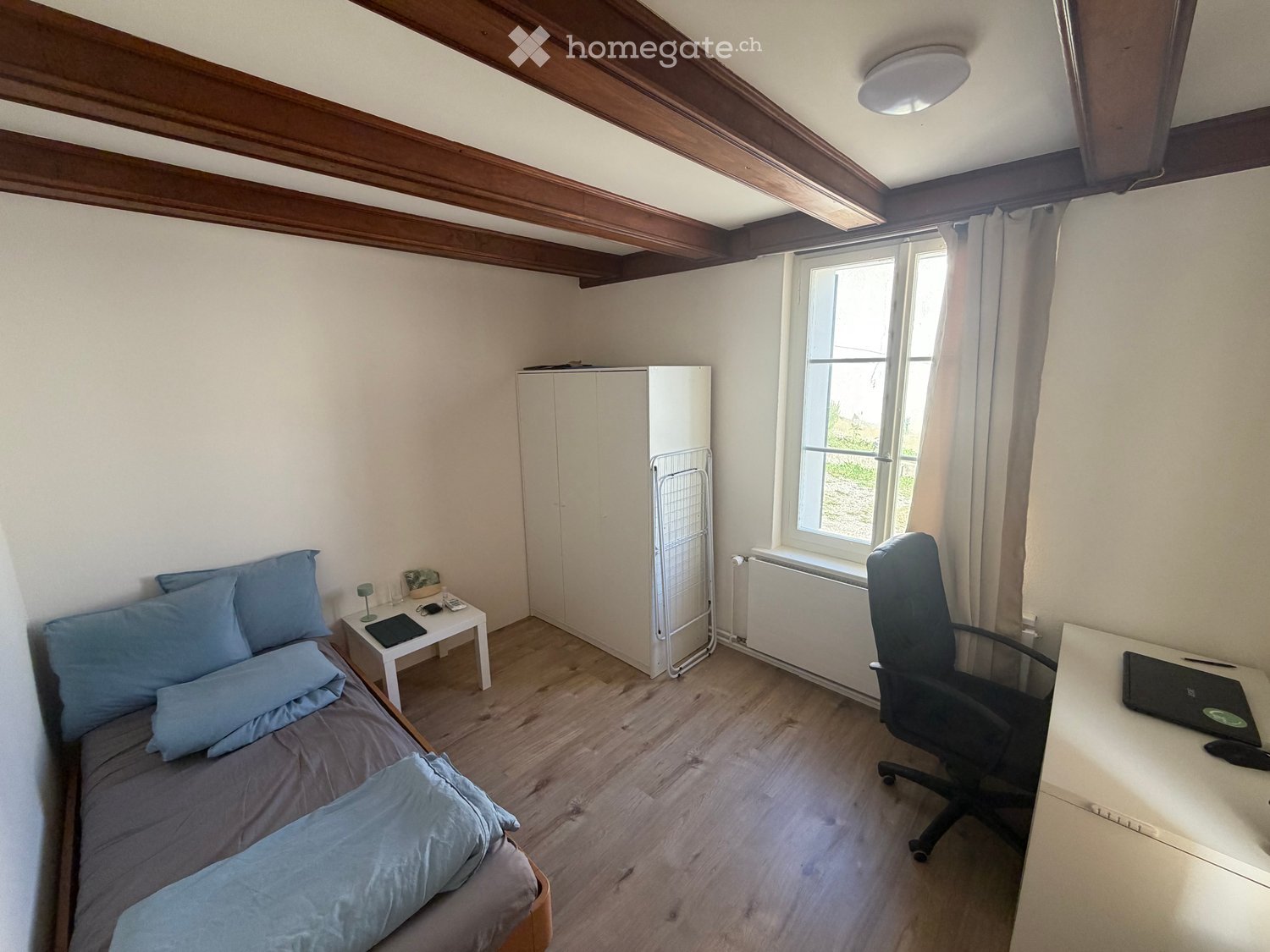 A small, well-lit bedroom with a single bed, a desk with a laptop, a chair, a small table with a lamp, and a white cabinet. The room features wooden flooring and exposed wooden beams on the ceiling.