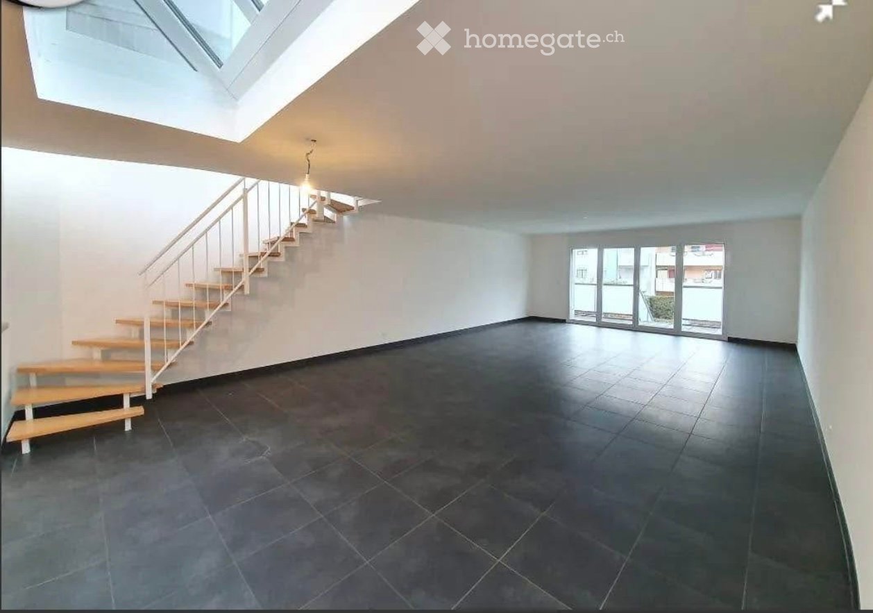Empty room with dark tiled floor, white walls, and a staircase leading to an upper level. Large window and skylight provide natural light.