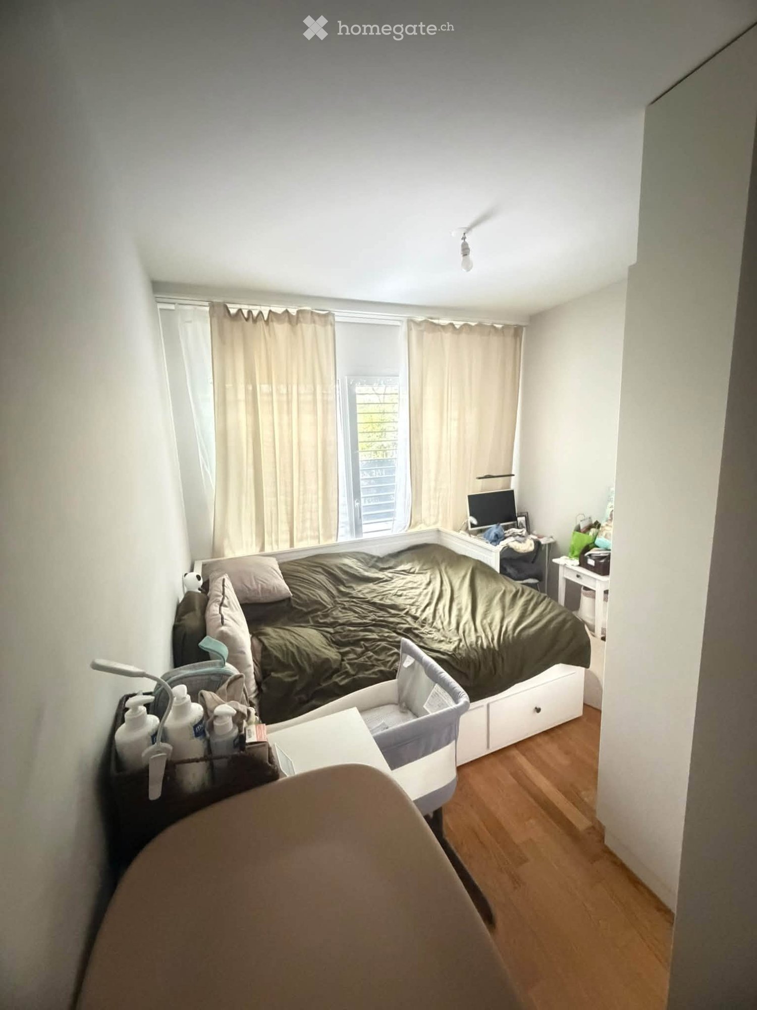 A small bedroom with a bed, baby cot, desk, and chair. The room has wooden flooring and large windows with curtains.