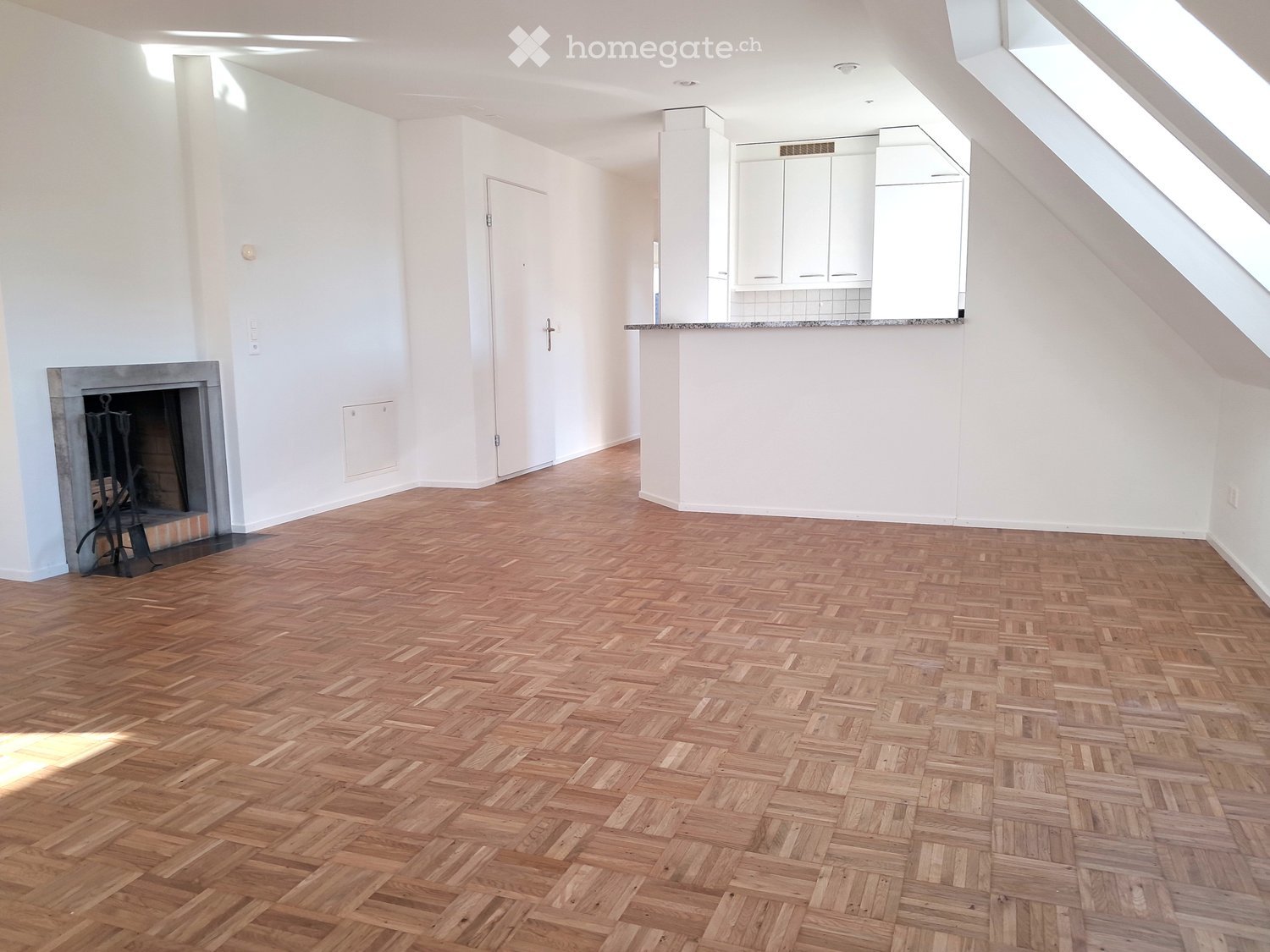Empty room with wooden herringbone flooring, white walls, fireplace, and a small kitchen area with cabinets and a countertop.